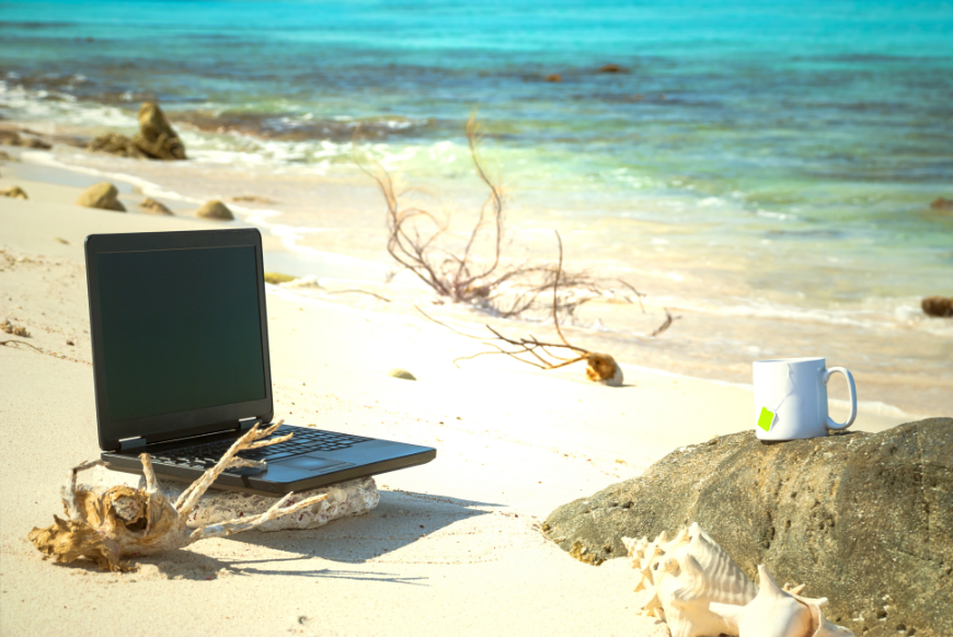 Retiring Abroad, Moving Abroad Image: A laptop and a coffee mug are sitting on a sandy beach.