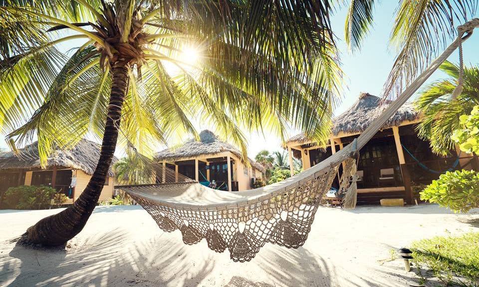 Vacation Time Image: A crocheted cream-colored beach hammock hovers effortlessly over white sand whilst affixed to two palm trees. Three thatched roof beach huts can be seen in the background.