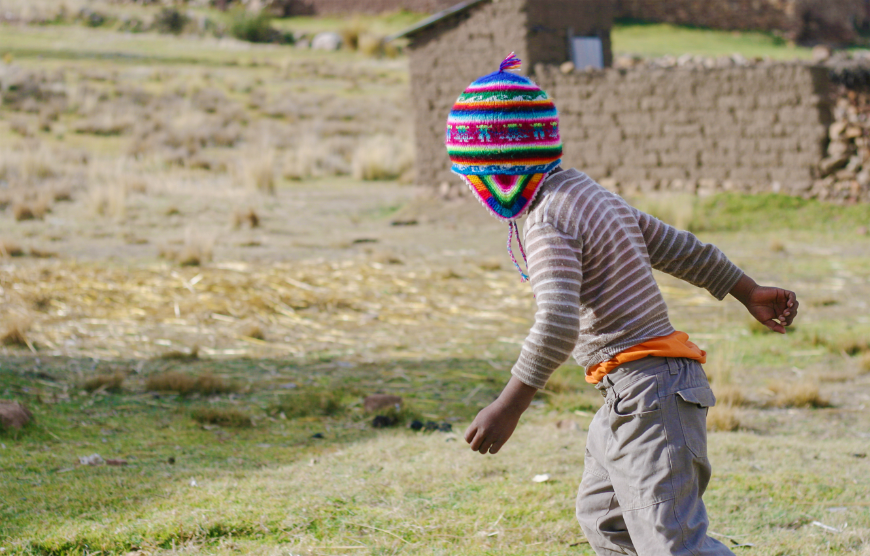 Peru Souvenir Image: A little boy wearing cargo pants, and tan and white striped shirt is running through a field whilst wearing a rainbow coloured chullo hat.