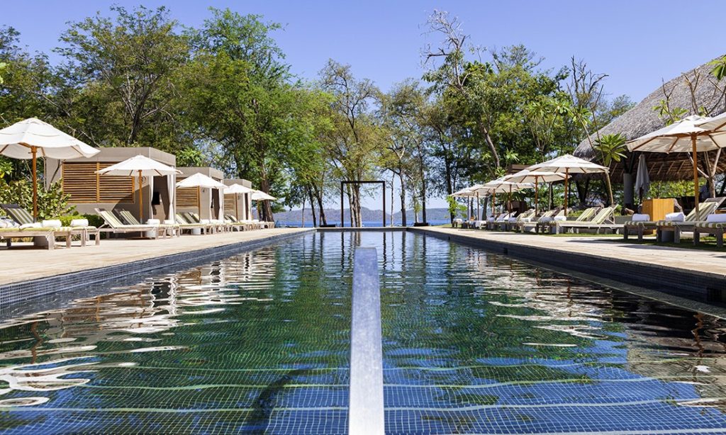 Unique Honeymoon Image: Lounge chairs, umbrellas, and cabanas flank a long pool looking out onto a beautiful view of trees, mountains, and ocean water.