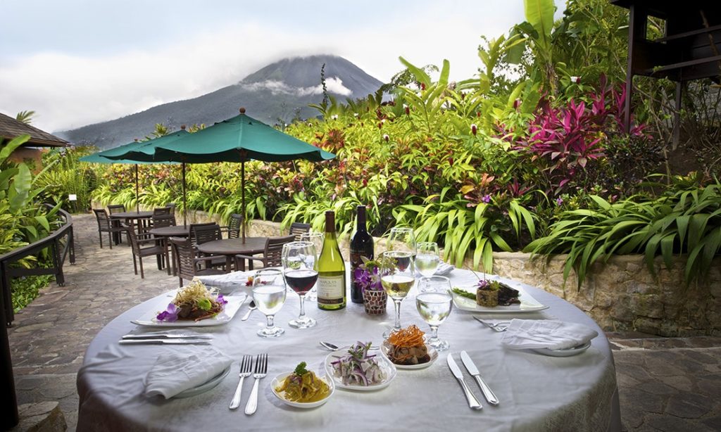 Unique Honeymoon Image: A delicious meal sits atop a white linen table setting, surrounded by lush and colorful foliage. A volcanic view is in the background.
