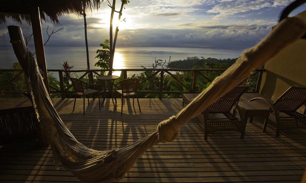 Unique Honeymoon Image: Views of a hammock, bistro set, and lounge chairs overlook a sloping rainforest canopy and sunset.