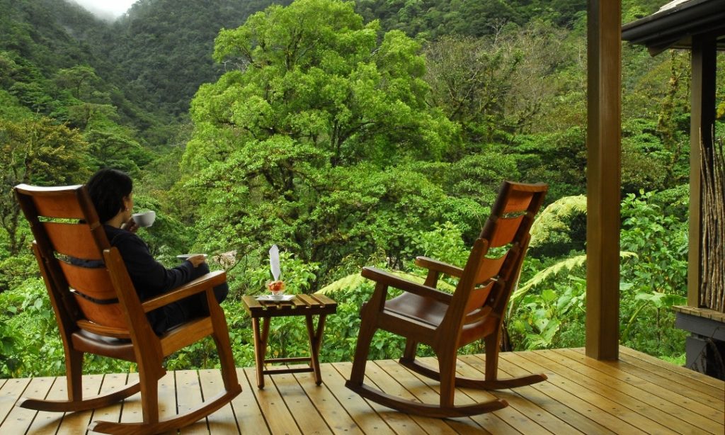 Unique Honeymoon Image: A woman sits in one of tea rocking chairs, sipping tea whilst quietly looking out at a lush green tropical forest. 