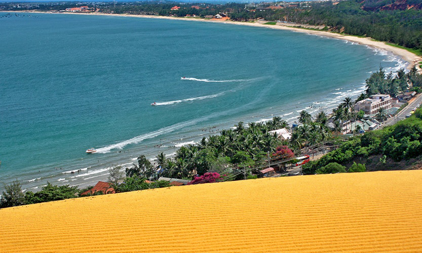 Vietnam Island Destination Image: Golden sand dunes overlook buildings, trees, and people playing and boating in the water.