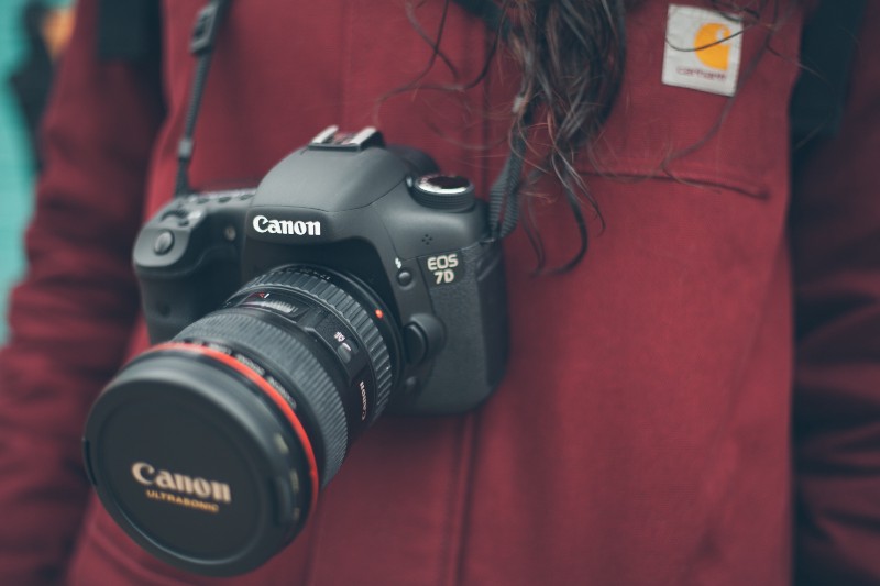 What To Pack Image: A woman (face not seen) with long brunette hair wears a red jacket and has a Canon camera hanging from her neck. 