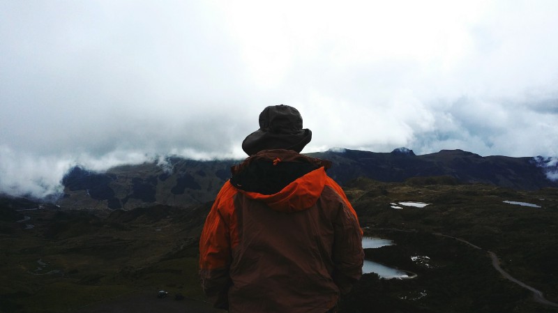 What To Pack Image: A person wearing a red hooded jacket and black hat (back to camera) stands gazing at a landscape of countryside, pools of water, and fog covered mountains.