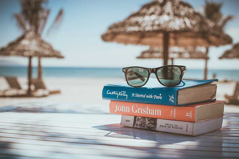 What To Pack Image: A stack of three books sits on a table; a pair of black sunglasses is perched on top of them. In the background is a sandy beach, and straw beach umbrellas.