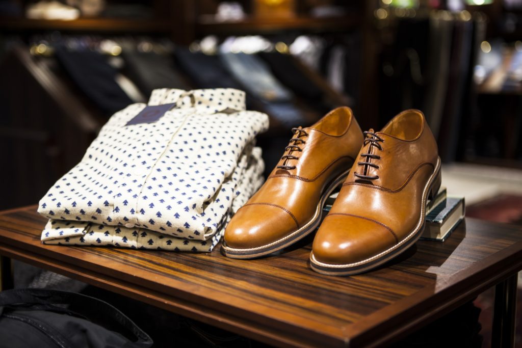 What To Pack Image: Polished light brown dress shoes, and a light-colored and patterned business casual buttonw-down dress shirt sit on a wooden table with books. 