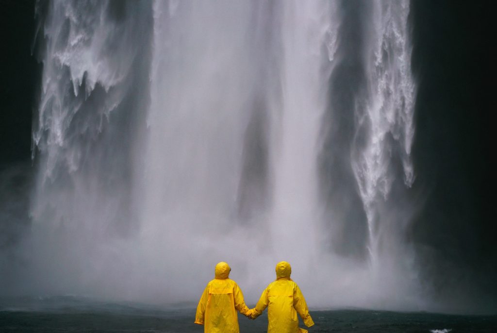 What To Pack Image: Two people with their backs to the camera hold hands whilst wearing yellow raincoats, and standing in front of / looking at a waterfall.
