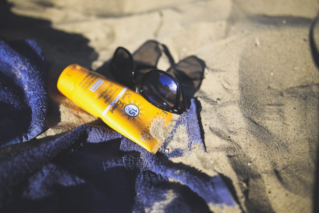 What To Pack Image: Black sunglasses and a yellow bottle of sun protectant cream rest upon a blue towel on a sandy beach.