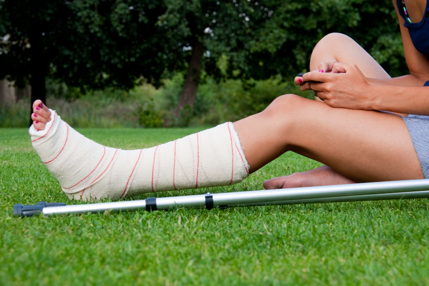Why Travel Insurance Is A Must Image: A woman sits in the grass using her phone. Her leg is broken and she has crutches.